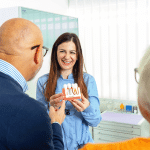 Female dentist holding anatomical model of teeth and explaining dental implant procedure to senior patients in clinic.